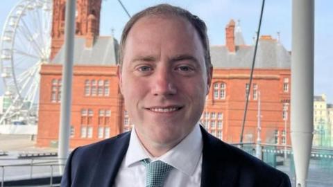 A head and shoulders picture of Rhys ab Owen, wearing a suit, shirt and tie, stood with Cardiff Bay's terracotta-glazed Pierhead building.