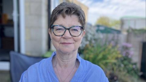 Karen Thompson stood in her garden. She is wearing a blue blouse and has glasses. Behind her there are some out of focus green bushes and pink flowers.