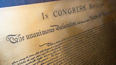 A piece of yellow, almost golden parchment bears the words of the Declaration of Independence. Written in block capital letters at the top of the page are the words: "IN CONGRESS. JULY 4, 1776".