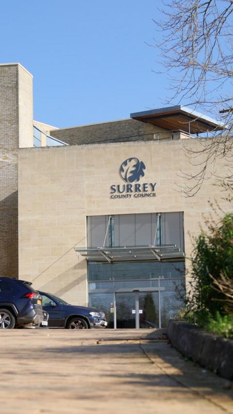 The outside of a light stone building. There is a sign for Surrey County Council above the glass entrance.