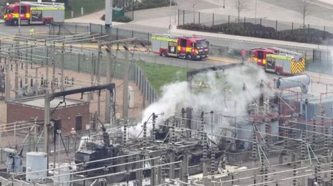 North Hyde substation on fire. Three fire engines are on a road next to it. Smoke is rising from the substation.