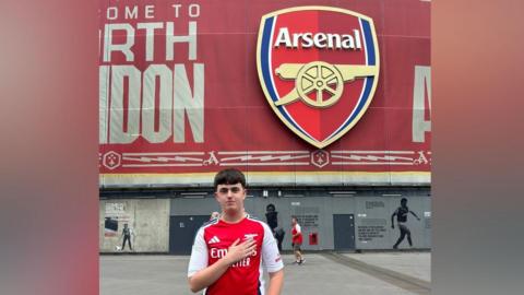 Alex had brown hair and is standing in front of the Emirates stadium which has a large red logo on it in the shape of a shield. It says Arsenal and has a large gold cannon on it. Alex is wearing a red and white Arsenal t-shirt and has his arm across his chest