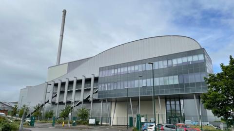 A large metal and glass incinerator with a tall grey chimney and cars and trees around it 