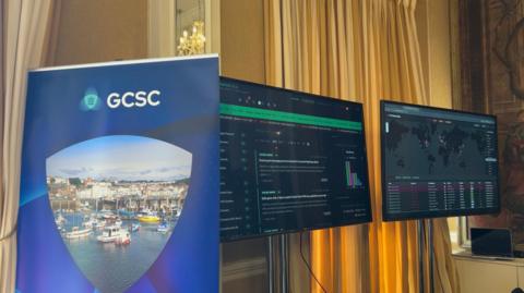 Two large black monitors on a yellow curtained wall show live tracking of cyber threats, including a map of the world. In front a blue banner which reads 'GCSC', with a photo St Peter Port Harbour 