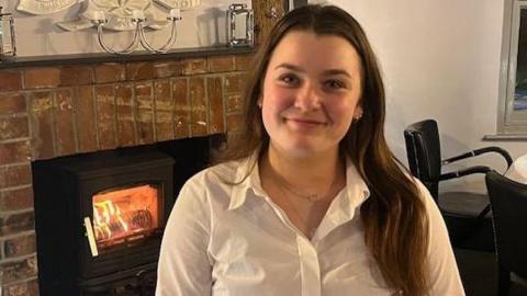 Willow is sitting wearing a white shirt in the pub she works in. She has long brown hair and a fireplace is visible behind her.