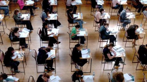 Five rows of school pupils sat at individual desks in a school hall taking exams. The camera is at a bird's-eye view angle. 