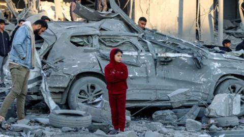 Men and children stand next to a destroyed car amidst debris and rubble by a collapsed building at the site of Israeli bombardment on a residential block in Jalaa Street in Gaza City on January 14, 2025