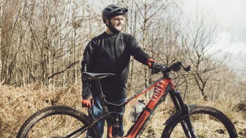 A man wearing an arm brace stands by his mountain bike in the woods.
