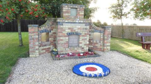 The brick memorial at the former RAF Metheringham airbase. Poppy wreaths are laying on the floor of the base, which is on a gravel floor.