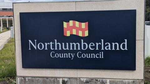 A navy blue sign in front of a building which reads 'Northumberland County Council'. The Northumberland flag made up of red and yellow squares is above the lettering.