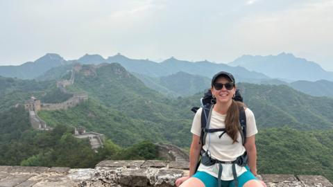 Ruth wears a white t-shirt and shorts and has a back pack on her ack.  She is sitting on part of the wall, which snakes through the hills in the background.