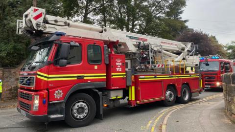 A stock image of a West Midlands Fire Service fire engine attending a different incident. The vehicle is parked on a road with another fire engine behind it. 