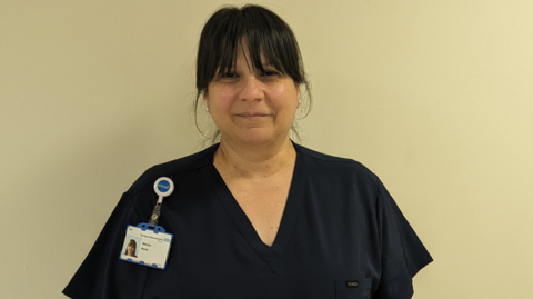 A woman with black hair wearing a navy V-neck medical scrubs with a security badge pinned to her right shoulder, she is standing in front of a white wall