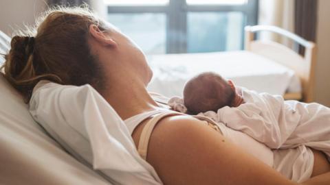 A mother rests in a hospital bed while gently holding her newborn baby.
