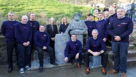 Members of the RNLI Whitby crew around the wire sculpture, wearing blue jumpers and black trousers and all smiling