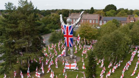 A large silver statue of a miner in Brownhills is adorned with a Union Jack Flag worn as a cape with St George's cross flags spread across the ground where the statue stands.