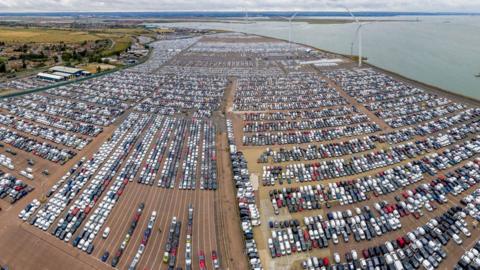 An aerial view of the port where thousands of cars are lined up. There are wind turbines on the coastline and the sea is visible to the right.