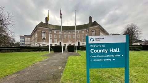 A sign pointing towards County Hall in Taunton, which is in the background. There are three flags, a green lawn and a path in front of the building. 