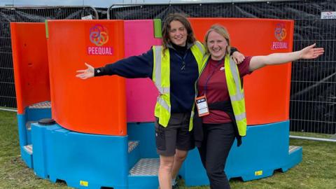 Two women stand with one arm around each other and the other outstretched to their side while smiling at the camera. They are both wearing yellow hi-vis vests and are standing in front of their design for a women's urinal. It is orange and blue in colour and has the word Peequal written on it 