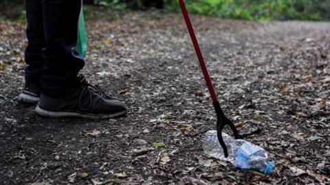 A generic photo of a person standing in a wooded area using a litter-picking tool to pick up a discarded water bottle. 