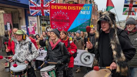 A group of protesters carrying signs through Exeter city centre that say things like 'women against the far right'. The front row are carrying drums. 