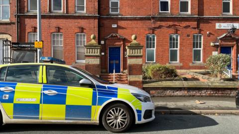 A marked police car parked up outside a redbrick building in front of a blue door