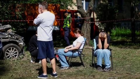 the image shows two women sat in fold out chairs with their heads in their hands and another man standing, facing away from the camera with his hands over his face. to their left is a burnt out car and in the background police tape and emergency workers.