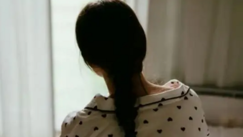 The back of a girl with long dark hair in a ponytail who is sitting looking out a window with white net curtains. She is wearing white pyjamas with black love hearts. 