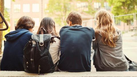 A group of young people sat with their back to the camera on a concrete bench. One is wearing a backpack they are wearing blue and grey colours. 