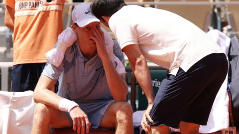 Jannik Sinner sits down in his seat and speaks to a trainer