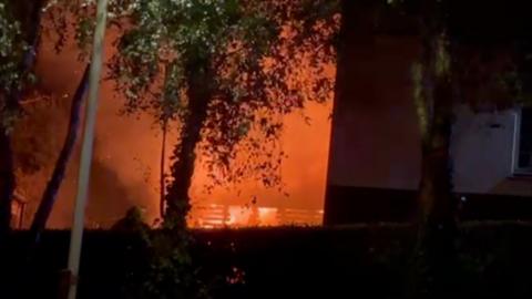 Large flames and thick orange smoke engulf an area behind several trees at night. A building is visible on the right side of the frame, partially silhouetted against the fire's glow, while dark foliage and a fence line the foreground. The scene is illuminated by the intense blaze, creating strong orange and black contrasts.