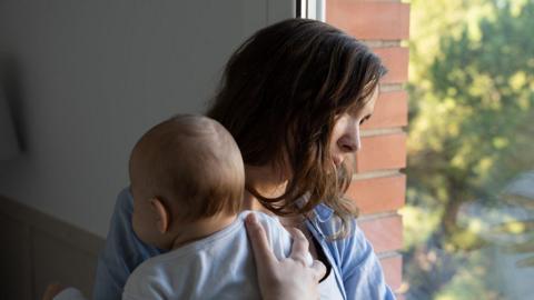 A woman holding a baby looks sadly out of a window.