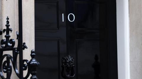 A black door in Downing Street with the Number 10 on it. A black railing can be seen in the foreground.