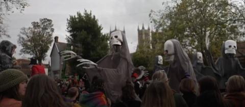 Three lanky skeletons with black capes on are towering above a crowd of people and in various dancing poses. Behind you can see Lincoln Cathedral in the distance.