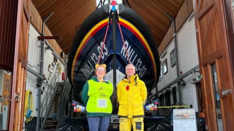 Caroline Densley, who is wearing blue leggings and a high-visibility vest, is standing next to a man in a yellow, all-weather suit. They are standing in front of an RLNI lifeboat.