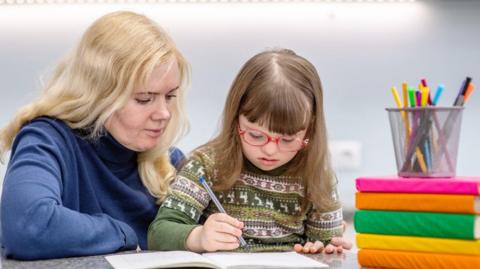 A young girl with downs syndrome is wearing red glasses and writing whilst a teacher is watching over and helping.