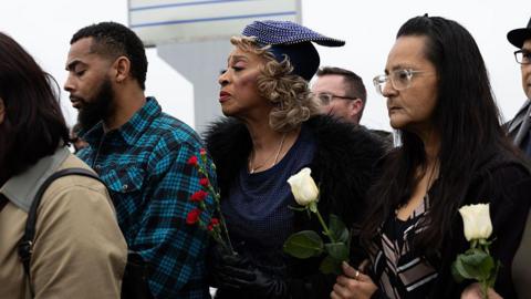 Man with goatee and black and teal checked shirt stands with eyes closed next to woman holding red carnations and wearing a blue dress and hat. Nearby, a woman with long brown hair and glasses holds a white rose. Another white rose, and other people's shoulders and parts of their faces can also be seen.