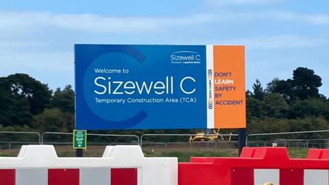 A Sizewell C board stands alongside a road. Red and white fences sit in front of it.