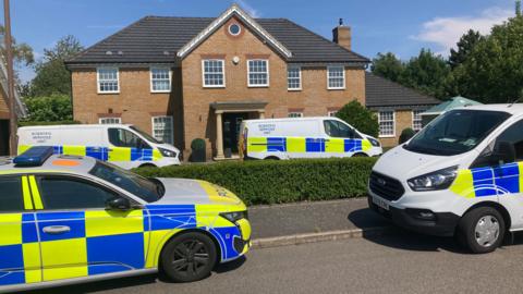 A police car and a few forensic vans are parked outside a large house, some on the street, some on the driveway