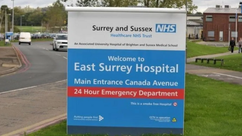 The entrance sign to East Surrey Hospital, with white lettering on a blue background, red brick buildings, roads, grassed areas and parked cars in the background.