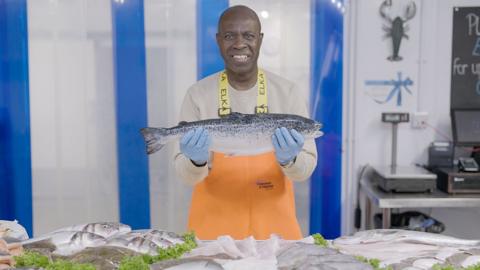 Clive Myrie holding a large oily fish in a fishmongers