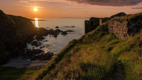 A grassy coastal path leads along a cliff overlooking a rocky cove at sunset. The sun hangs low over the sea, casting golden light across the water and surrounding hillsides. Stone walls and ruins stand on the right, while dark rocks line the shoreline below beneath a sky streaked with clouds.