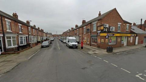 Image of a street containing terraced houses, with a convenience store on the near right hand corner and parked vehicles on both sides of the road 