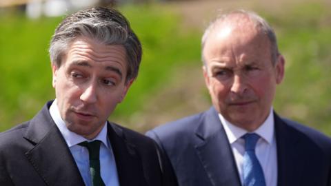 Simon Harris and Micheál Martin look to the left of the camera. They are both in dark suits and look deep in thought.