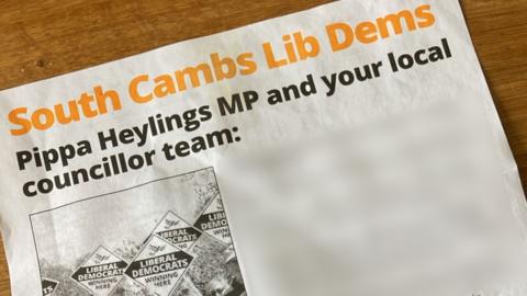 A leaflet titled 'South Cambs Lib Dems', with a sub heading that says 'Pippa Heylings MP and your local councillor team'. The headline is in yellow and stretches across the width of the page. Below the sub heading, which is in black typeface, is a black and white photograph, which shows diamond-shaped Liberal Democrats signs being held in the air. A section of writing to the right of the image has been blurred out, so cannot be read.