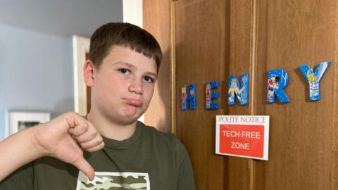 A young boy holds his thumb downwards, outside his bedroom door, which has a sign 'Polite notice: Tech free zone'