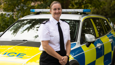 A woman in a police uniform is standing by a police car with her hands folded in front of her. Her hair is tied back and she is smiling at the camera.