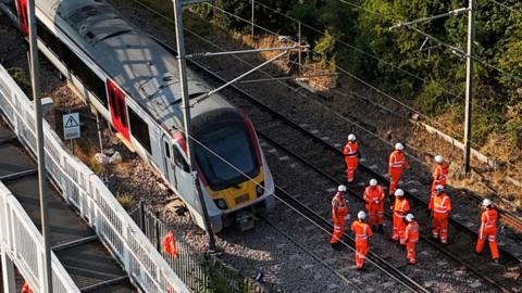 Part of a Greater Anglia train service derailed at Wickford in Essex. A number of people wearing fluorescent orange outfits with white helmets are stood on the tracks near the train.