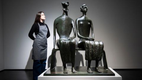 A bronze sculpture of a couple sitting on a bench. A woman in a black top and grey apron stands next to the sculpture with her hands behind her back, looking at it.
