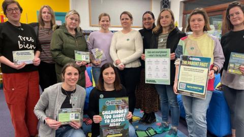 A large group of women stand together holding up posters, stickers and leaflets. They are standing in a brightly coloured library.